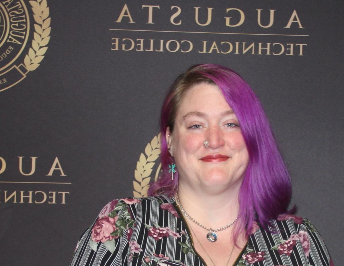 Alina Walls, a caucasian female with lavendar hair stands in front of a background with the ATC seal and name in gold and black. She is pictured wearing a black and gold patterned top.